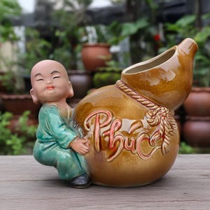 May include: A ceramic figurine of a bald child wearing a green tunic, leaning against a large, brown, gourd-shaped vessel. The vessel features a sculpted rope detail and the word "Phúc" embossed in red lettering on its rounded side.
