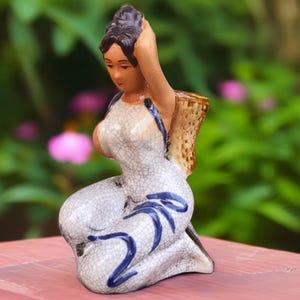 Könnte beinhalten: Keramikfigur einer knienden Frau mit hochgestecktem Haar und einem Korb auf dem Rücken. Die Figur ist mit einer weißen Craquelé-Glasur und blauen Akzenten bemalt. Die Frau trägt ein Kleid mit blauen Verzierungen.