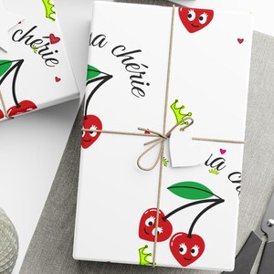Puede incluir: Papel de regalo blanco con un diseño de cereza roja y el texto "Ma Cherie" en negro. La cereza tiene un tallo y hojas verdes y una cara linda. También hay pequeños corazones rojos y coronas verdes en el papel de regalo.