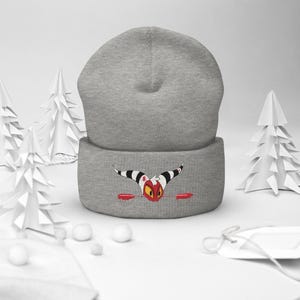 Op de afbeelding: Grijze beanie met een rood en wit cartoonfiguur geborduurd op de voorkant. Het figuur heeft hoorns en kijkt omhoog.