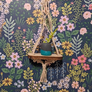 Könnte beinhalten: Ein kleiner, handgefertigter Makramee-Pflanzenaufhänger mit einem Holzregal, das einen kleinen, dunkelgrauen Topf mit türkisfarbenem Boden und einer grünen Pflanze hält. Der Hintergrund ist eine florale Tapete in Marineblau mit rosa, gelben und weißen Blumen.