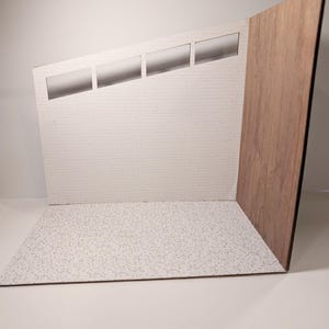 May include: A miniature studio set with a white brick wall, a wood-grain panel, and a speckled floor. The wall has three rectangular cutouts. The set is designed for product photography or small-scale displays.