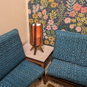 Puede incluir: Dos sillones de tela azul con marcos de madera y una mesa auxiliar de madera con una lámpara. La lámpara tiene una base de madera y una pantalla cilíndrica roja y blanca. El fondo es un papel tapiz floral con flores rosas, amarillas y blancas sobre un fondo azul.