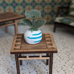 May include: A small wooden table with a square top and a decorative pattern. A white planter with blue stripes sits on the table, holding a small green plant with three leaves.