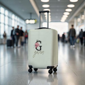 Könnte beinhalten: Ein cremefarbener Rollkoffer mit floralem Design und dem Aufdruck "gwen". Das Design beinhaltet eine rosa Blume und den Buchstaben "G". Der Koffer hat einen ausziehbaren Griff und vier schwarze Räder. Der Hintergrund ist ein verschwommenes Flughafenterminal.