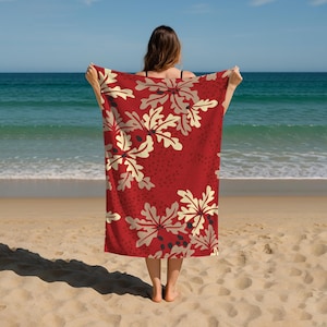 May include: A red beach towel with a floral design. The towel displays large, cream-coloured flowers with dark blue accents. The background is a solid red with a subtle dotted texture. The towel is held up on a sandy beach with the sea in the background.
