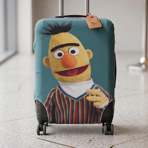 K&ouml;nnte beinhalten: Eine t&uuml;rkisfarbene Kofferh&uuml;lle mit einer Cartoonfigur mit gelber Haut, einer orangefarbenen Nase und einem gestreiften Hemd. Die Figur l&auml;chelt mit offenem Mund. Das Gep&auml;ck hat ein Gep&auml;ckanh&auml;nger.