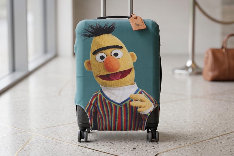 K&ouml;nnte beinhalten: Eine t&uuml;rkisfarbene Kofferh&uuml;lle mit einer Cartoonfigur mit gelber Haut, einer orangefarbenen Nase und einem gestreiften Hemd. Die Figur l&auml;chelt mit offenem Mund. Das Gep&auml;ck hat ein Gep&auml;ckanh&auml;nger.