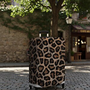 Pode incluir: Uma mala com rodas com um design de estampa de leopardo. A mala tem uma al&ccedil;a e rodas pretas. O fundo mostra uma rua de paralelep&iacute;pedos, uma &aacute;rvore e um edif&iacute;cio com um caf&eacute;.