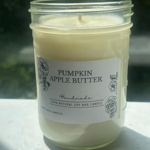 May include: A clear glass jar candle filled with off-white soy wax and a single centered wick. The white label features the text PUMPKIN APPLE BUTTER, Handmade, 100% NATURAL SOY WAX CANDLE, and WISPY WICK CANDLES, decorated with black floral line art.