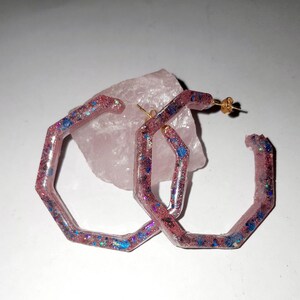 Op de afbeelding: Een paar roze zeshoekige creolen met iriserende glitter ingebed in de hars. De oorbellen hebben gouden oorbelsluitingen.