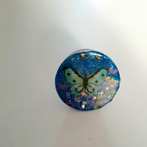 Peut inclure: Un objet décoratif rond bleu avec un motif de papillon. Le papillon est vert clair avec des détails noirs. L'objet est orné de paillettes et de petits accents d'étoiles. Le fond est un dégradé de bleu et de blanc.