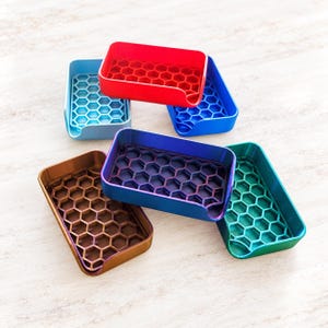 May include: A collection of five rectangular soap dishes in red, light blue, dark blue, bronze, and teal. Each dish features a raised internal honeycomb grid pattern and a small circular cutout on one of the shorter sides for drainage.