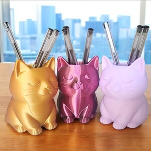 May include: Three cat-shaped pen holders in metallic gold, metallic magenta, and matte lavender. Each holder features a stylized feline face with closed eyes and whiskers, designed to store pens or pencils upright in the open top cavity.