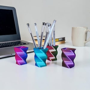 May include: A collection of four 3D-printed pen holders in a twisted design, each with a unique color gradient. The holders are filled with clear pens and sit on a white desk, alongside a laptop and a coffee mug. The holders are approximately 4 inches tall.