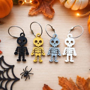 May include: Four articulated skeleton keychains in black, yellow, gray, and white. Each figure features a round skull, a rib cage, and jointed limbs, attached to a circular metal wire ring with a screw-style closure for securing keys.