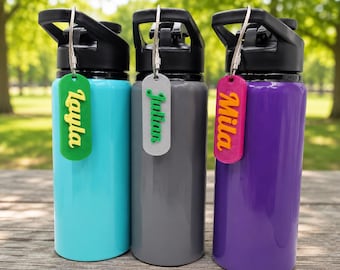 May include: Three stainless steel water bottles with black lids, each featuring a personalized rectangular tag attached by a silver wire. The tags are green with yellow text reading Layla, gray with green text reading Julian, and pink with orange text reading Mila.