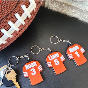 May include: Three orange and black football jersey keychains with white names and numbers. The keychains are "Noah 3", "Liam 5", and "Mason 1".
