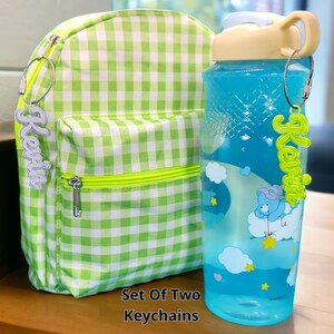May include: Set of two green and white gingham backpacks with matching keychains. The keychains feature the names "Kevin" and "Karina" in white letters.