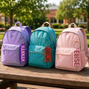 May include: Three backpacks in light purple, teal, and light pink, each featuring a front zippered pocket and a side mesh pocket. Attached to the top handle of each bag is a silver ring with a vertical rectangular name tag reading HANNAH, JACK, or ELISE.