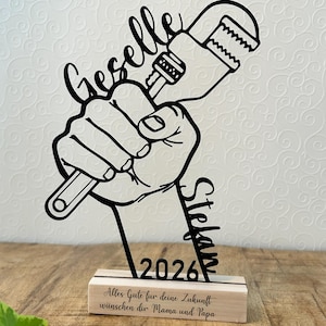 Könnte beinhalten: Schwarze Metallskulptur einer Hand, die einen Schraubenschlüssel hält, mit den Namen "Geselle" und "Stefan" und dem Jahr "2026". Die Skulptur ist auf einem Holzsockel mit deutschem Text montiert.
