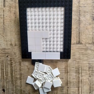 May include: A rectangular building block baseplate with a black border and a white studded center. Several white square tiles are attached to the baseplate, and a small pile of loose white square tiles is positioned directly below the baseplate.