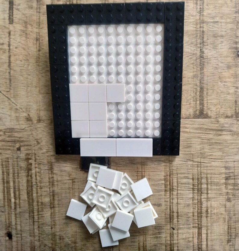 May include: A rectangular building block baseplate with a black border and a white studded center. Several white square tiles are attached to the baseplate, and a small pile of loose white square tiles is positioned directly below the baseplate.