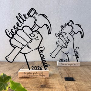 Könnte beinhalten: Zwei schwarze Schreibtischdekorationen mit Hammer-Motiv. Jede zeigt einen Hammer in einer Hand, mit einem Namen und dem Jahr 2026. Auf einer steht "Geselle Dominik" und auf der anderen "Geselling Sonja". Eine Basis lautet "Herzlichen Glückwunsch zu deiner bestandenen Prüfung".