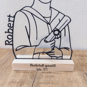May include: A black metal graduation figure with the name "Robert" on the side. The figure holds a diploma and is mounted on a wooden base with the text "Meisterhaft gemacht! K&ouml;ln 2025".