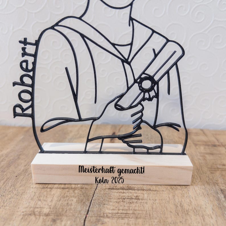May include: A black metal graduation figure with the name "Robert" on the side. The figure holds a diploma and is mounted on a wooden base with the text "Meisterhaft gemacht! K&ouml;ln 2025".