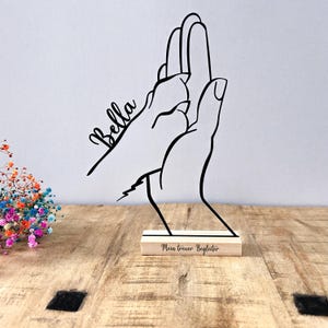 May include: A black metal sculpture of a hand giving a paw a high five. The name "Bella" is written in cursive on the paw. The sculpture is mounted on a wooden base with the text "Mein treuer Begleiter".