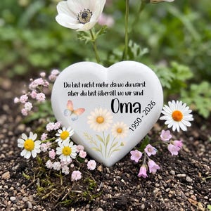 Puede incluir: Una piedra conmemorativa blanca en forma de corazón con el texto en alemán "Du bist nicht mehr da wo du warst aber du bist überall wo wir sind", la palabra "Oma", los años "1950 - 2026", una ilustración de mariposa y margaritas.