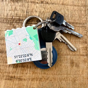 May include: A keychain with a map design, featuring a red heart marker and coordinates 51°22'22.6"N 6°30'32.9"E. The keychain is attached to a metal ring with several keys. The background is a wooden surface.