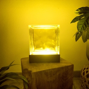 Può includere: Una lampada a blocchi di vetro trasparente illuminata da una calda luce gialla. La lampada è posizionata su una base scura e posta su un tavolino di legno. Lo sfondo è di colore giallo pieno, con una pianta in vaso a destra.