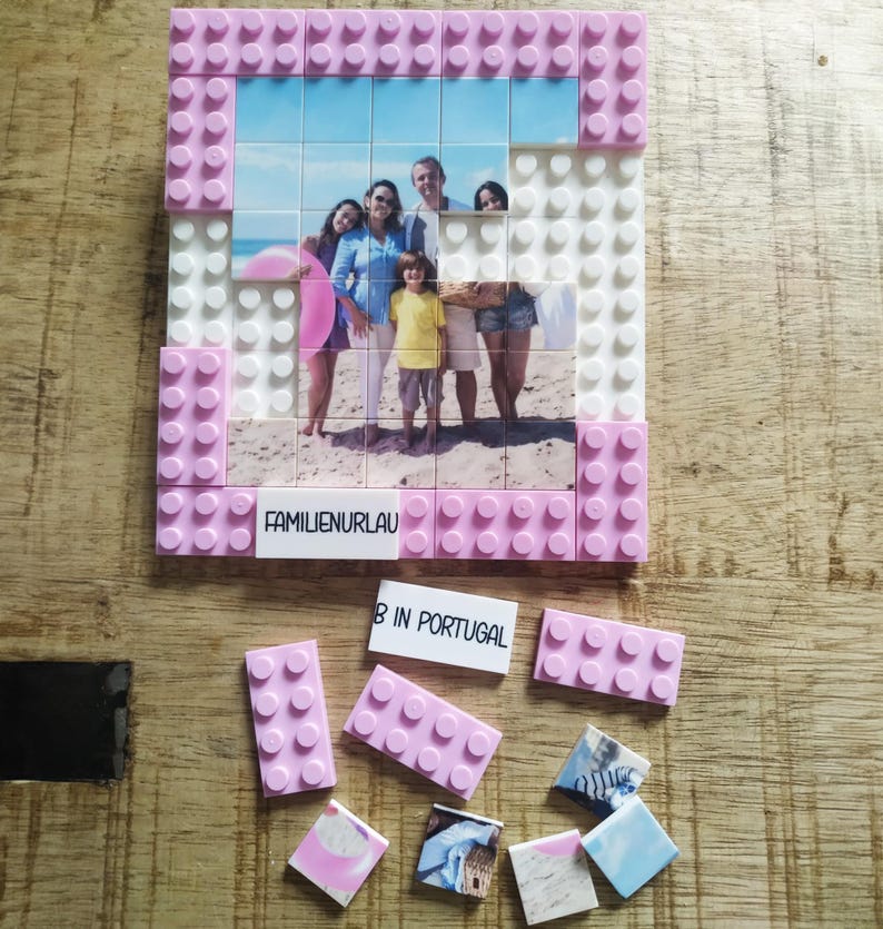 May include: A custom photo mosaic constructed from interlocking plastic building blocks. The frame is made of pink and white bricks. The central image shows a family on a beach. A white brick features the text FAMILIENURLAUB, with additional loose bricks and photo tiles labeled B IN PORTUGAL.