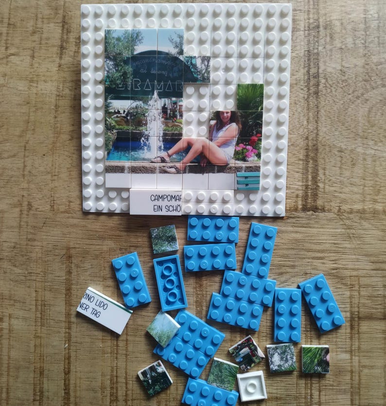 May include: A white plastic building block baseplate featuring a photo mosaic of a fountain and a person sitting on a ledge. The text CAMPOMARINO LIDO EIN SCH&Ouml;NER TAG is printed on a tile. Scattered blue building blocks and additional photo tiles.