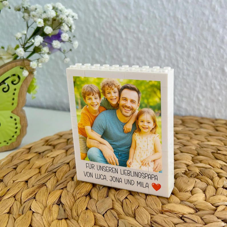 May include: A white block photo frame featuring a family portrait of a father and three children. The frame has the text "FUR UNSEREN LIEBLINGSPAPA VON LUCA, JONA UND MILA" and a red heart. The frame is on a woven surface.