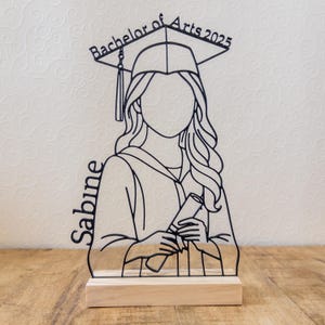 May include: A black metal graduation figure with the text "Bachelor of Arts 2025" and the name "Sabine". The figure is holding a diploma and wearing a graduation cap. It is mounted on a wooden base.