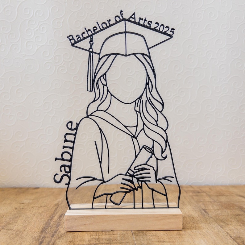 May include: A black metal graduation figure with the text "Bachelor of Arts 2025" and the name "Sabine". The figure is holding a diploma and wearing a graduation cap. It is mounted on a wooden base.