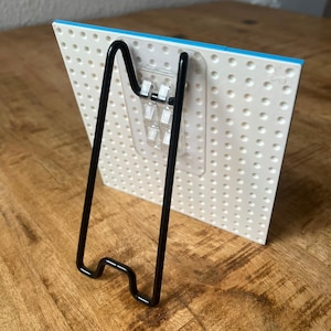 May include: A white plastic building block baseplate with a grid of circular studs, featuring a clear adhesive hook attached to the back. A black metal wire easel stand is hooked into the adhesive mount to support the baseplate in an upright position.
