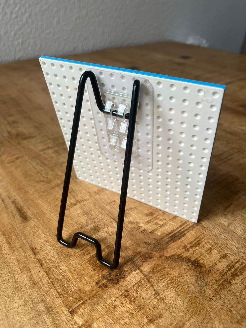 May include: A white plastic building block baseplate with a grid of circular studs, featuring a clear adhesive hook attached to the back. A black metal wire easel stand is hooked into the adhesive mount to support the baseplate in an upright position.