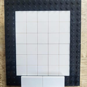 May include: A rectangular frame constructed from black plastic building bricks with a central grid of smooth white square tiles. The border consists of black studs, while the interior is filled with a flat, white surface made of square pieces.