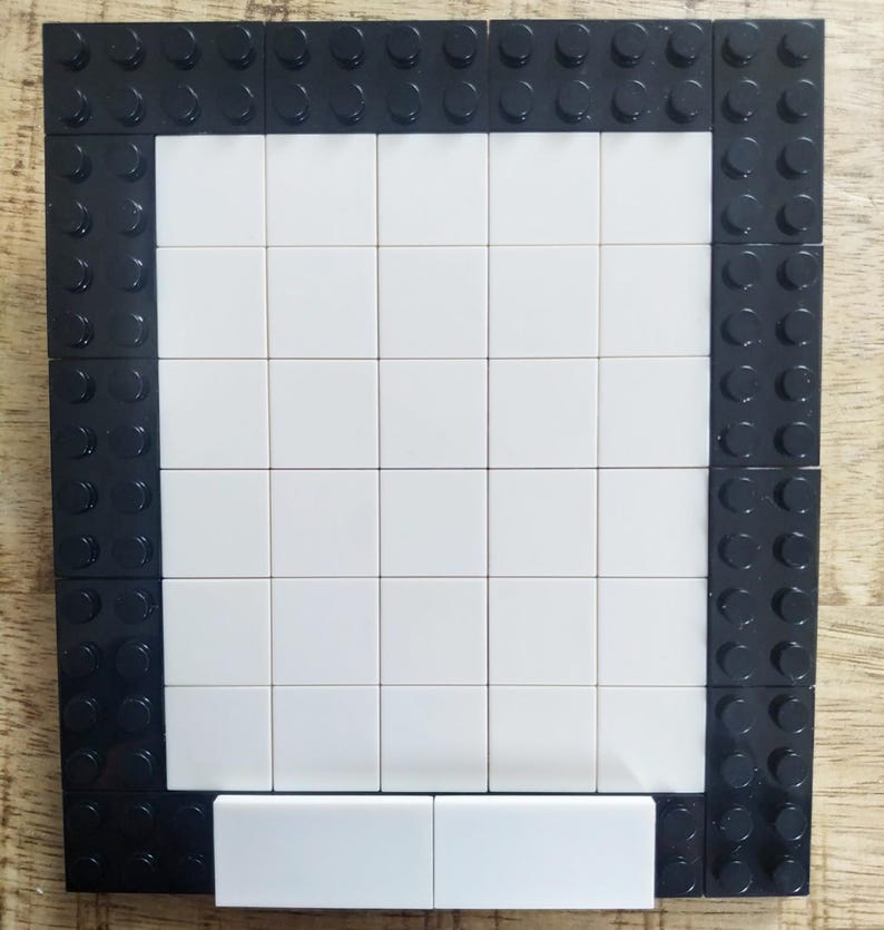May include: A rectangular frame constructed from black plastic building bricks with a central grid of smooth white square tiles. The border consists of black studs, while the interior is filled with a flat, white surface made of square pieces.