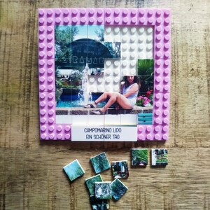 May include: A square building block frame in light pink featuring a photo mosaic of a person sitting by a fountain. The text CAMPOMARINO LIDO EIN SCH&Ouml;NER TAG is printed on a white tile at the bottom. Several loose photo tiles lie below the frame.