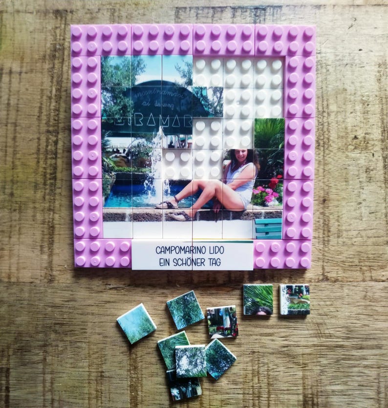 May include: A square building block frame in light pink featuring a photo mosaic of a person sitting by a fountain. The text CAMPOMARINO LIDO EIN SCH&Ouml;NER TAG is printed on a white tile at the bottom. Several loose photo tiles lie below the frame.