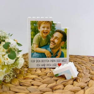 May include: A white block photo frame featuring a picture of a smiling father and child. The frame has the text "F&Uuml;R DEN BESTEN PAPA DER WELT ICH HAB DICH LIEB" with a red heart. The frame is on a woven surface.