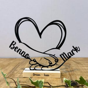 Op de afbeelding: Zwarte metalen sculptuur met een hart boven in elkaar grijpende handen. De namen "Benno" en "Mark" staan aan weerszijden. De basis is een houten blok met de tekst "Dream - Team".