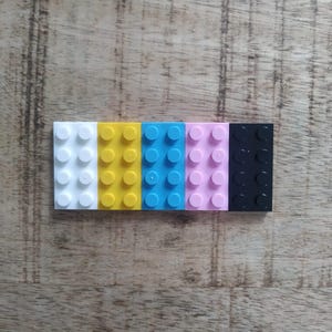 May include: A row of five rectangular plastic building bricks, each featuring eight circular studs on the top surface. The bricks are arranged side-by-side in a sequence of white, yellow, bright blue, light pink, and black colors.