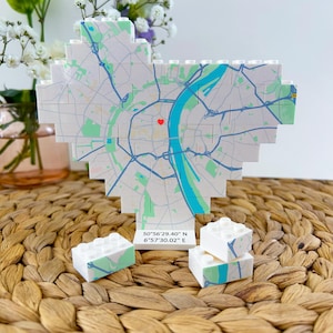 Op de afbeelding: Een hartvormige kaart gemaakt van witte blokken, met een stadskaart met blauwe en groene accenten. Een rood hart markeert een locatie. Twee kleinere bloksets staan ervoor. Coördinaten zijn afgedrukt.