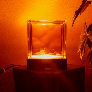 May include: A clear glass block lamp illuminated with a warm, orange glow. The lamp sits on a dark, square base, casting a soft light against an orange wall. The overall aesthetic is minimalist and modern.